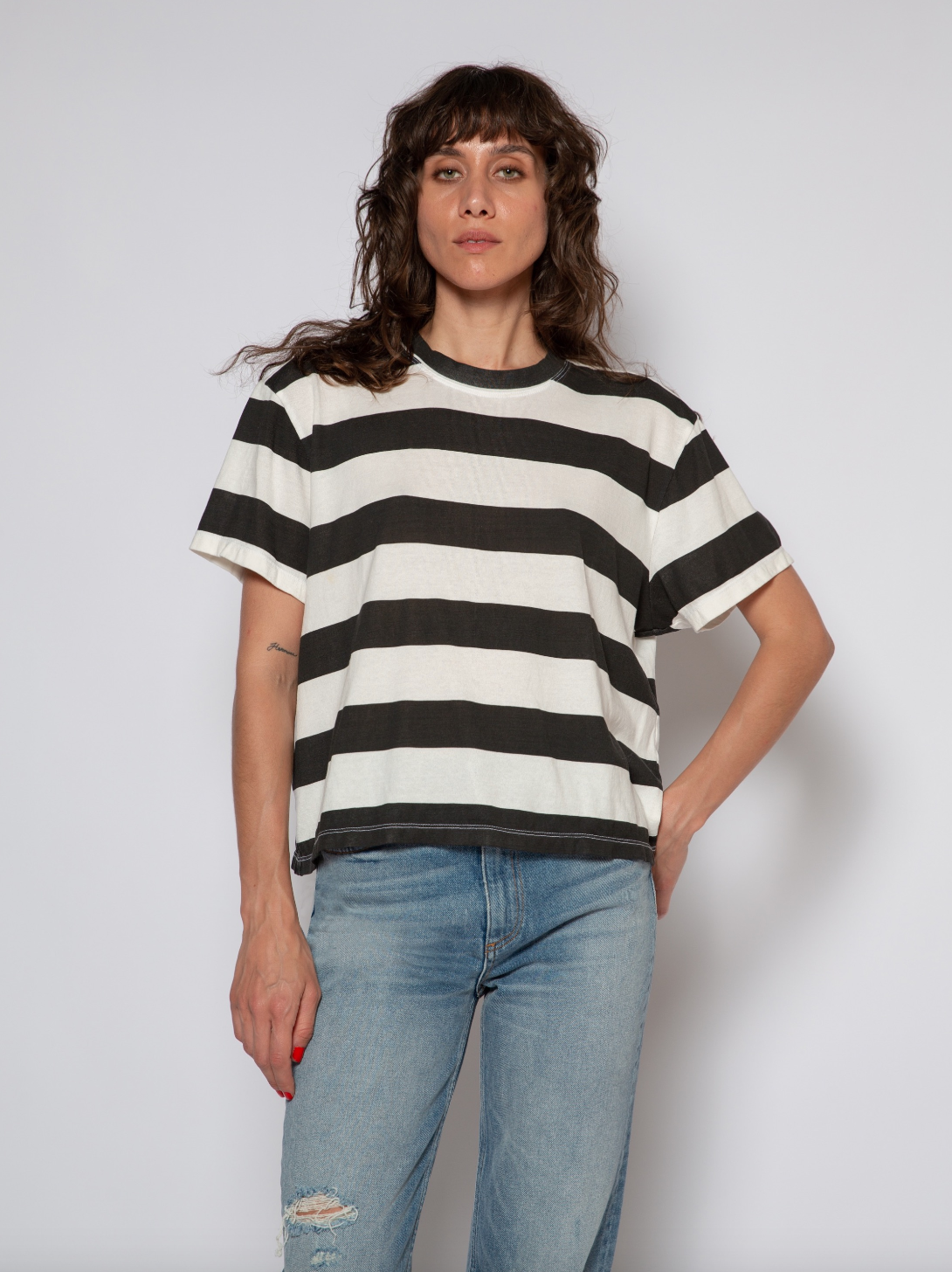 A woman with curly shoulder-length hair wearing a black and white striped boy tee from ASKK and blue jeans stands against a plain background in Scottsdale, Arizona, looking at the camera.