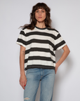 A woman with curly shoulder-length hair wearing a black and white striped boy tee from ASKK and blue jeans stands against a plain background in Scottsdale, Arizona, looking at the camera.