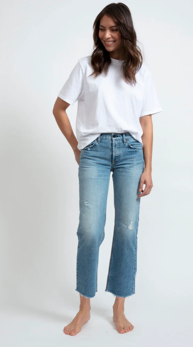 A woman in a white t-shirt and distressed blue jeans stands barefoot, smiling subtly and placing one hand in her pocket, exuding ASKK style with the Berkeley Low Rise Straight jeans.