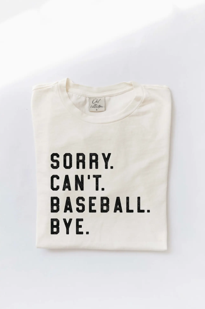 A folded Sorry. Can't. Baseball. Bye. Mineral Washed Graphic Top Cream from Oat Collective, with the text printed in bold, black letters on the front, photographed against a plain white background in a Scottsdale, Arizona bungalow.
