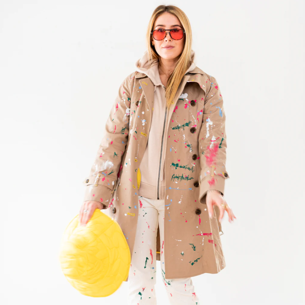 A woman in a paint-splattered KR Trench by Kerri Rosenthal and sunglasses holds a yellow hard hat, standing against a white background in Scottsdale, Arizona.