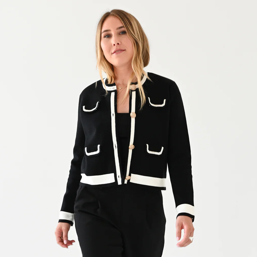 A woman models a stylish black and white Beau-Lero Cardigan Sailor Black jacket with Arizona-style trim details, paired with black trousers, standing against a white background. (Brand: Kerri Rosenthal)