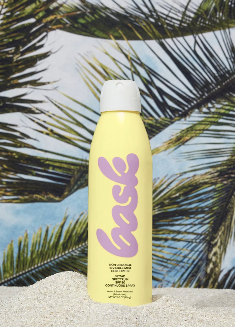 A can of Faire Bask Sunscreen SPF 50 labeled "face" in purple letters on a yellow background, positioned on sand with palm fronds in the background near an Arizona bungalow.