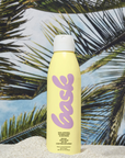 A can of Faire Bask Sunscreen SPF 50 labeled "face" in purple letters on a yellow background, positioned on sand with palm fronds in the background near an Arizona bungalow.