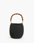 The Pot de Miel by Clare Vivier is a round, black basket bag featuring a brown bamboo handle. Its handwoven straw texture adds depth to its simple, elegant design against a white background.