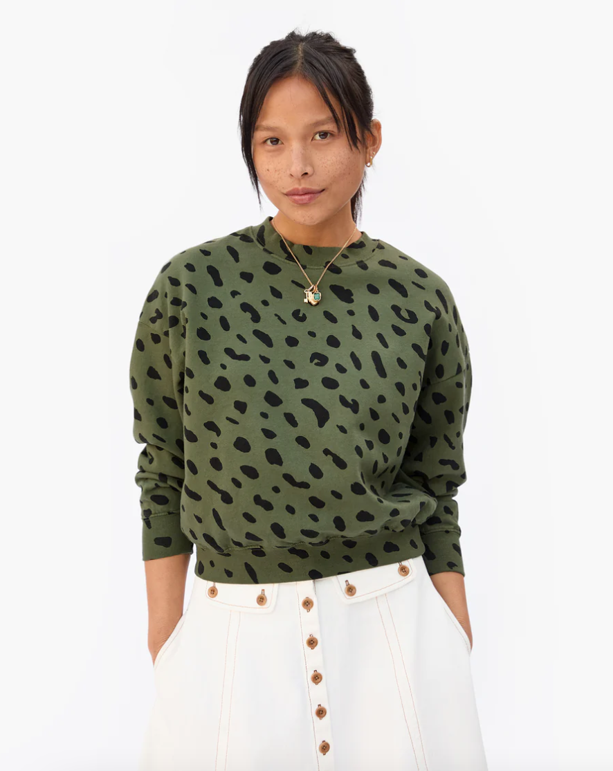 A woman in a green leopard print sweater and white buttoned skirt, standing against a plain background, looks at the camera. She wears a subtle Clare Vivier necklace.