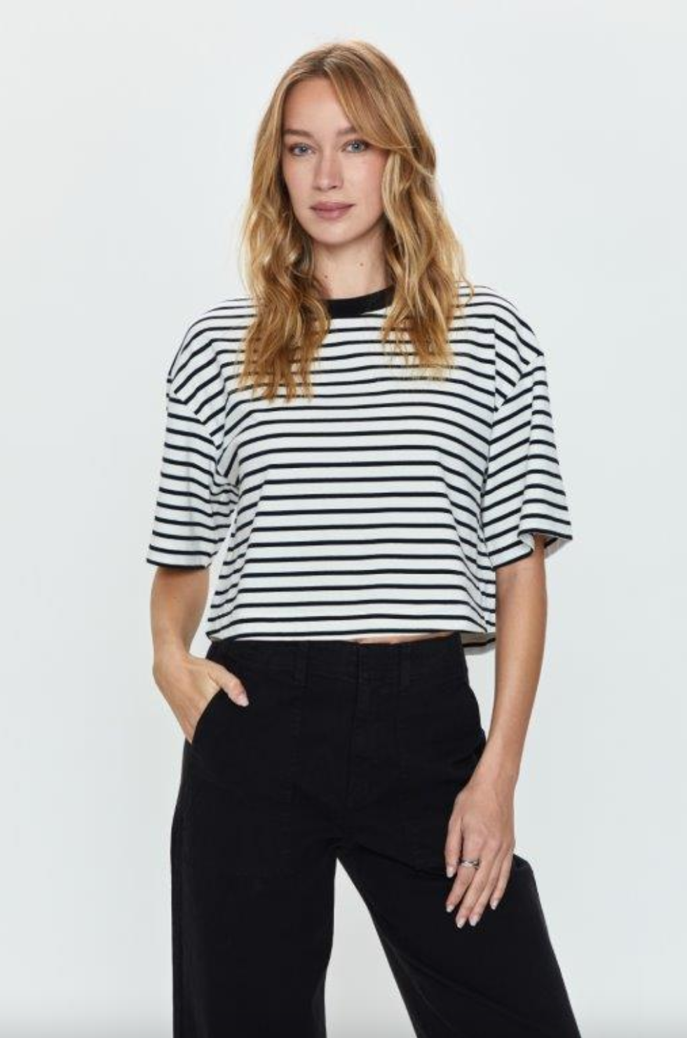 A woman wearing a Pistola Mae Cropped Tee, a Los Angeles-based black and white striped T-shirt, and black pants, standing against a white background, with her hand on her hip and looking directly at the camera.
