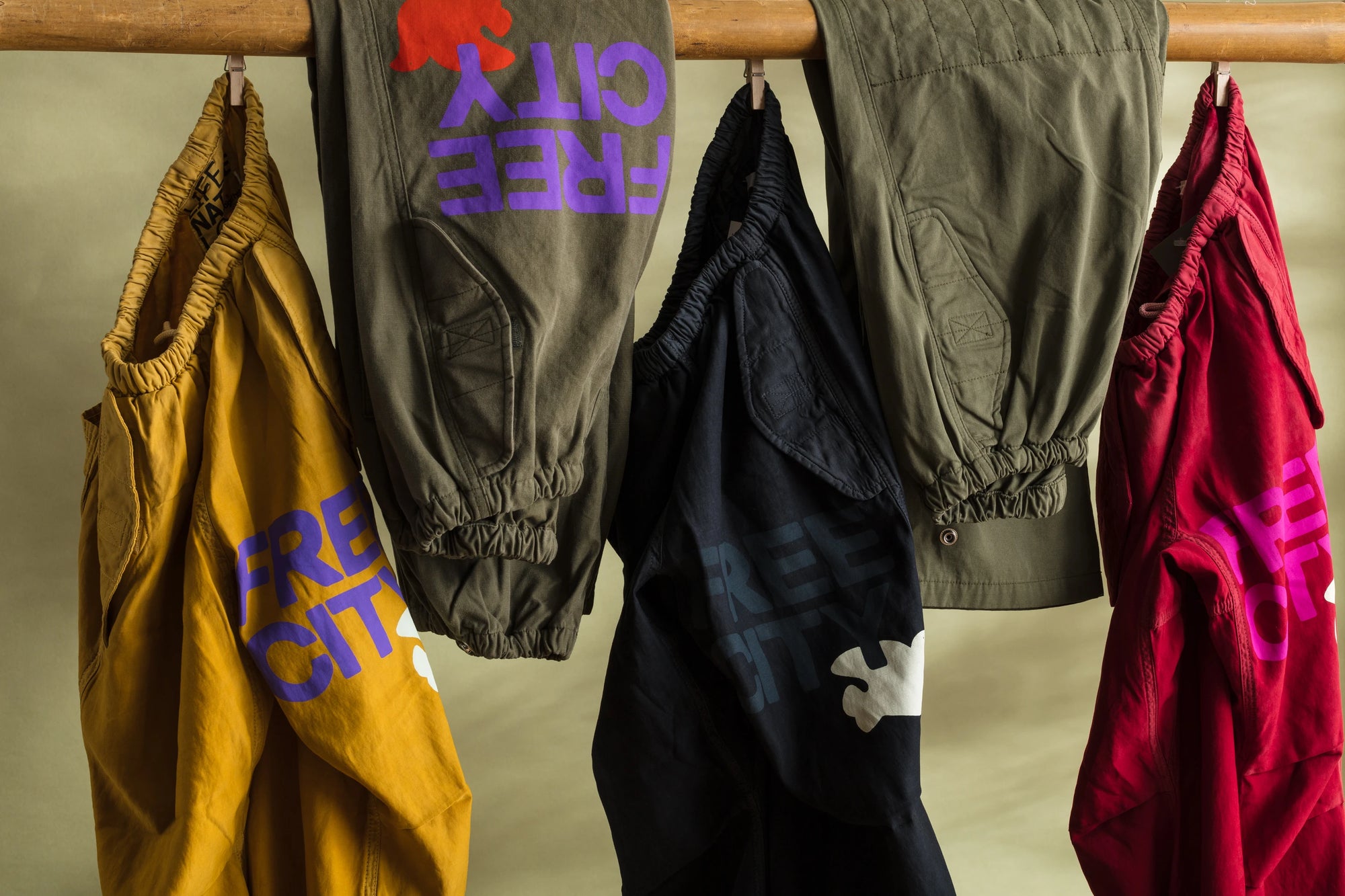 A line of sweatshirts and hoodies in various colors, including pink, green, orange, gray, and beige, hang from a rope on metal hangers. Each garment has a "FREE CITY" logo and a bird graphic. The background is a plain white wall.