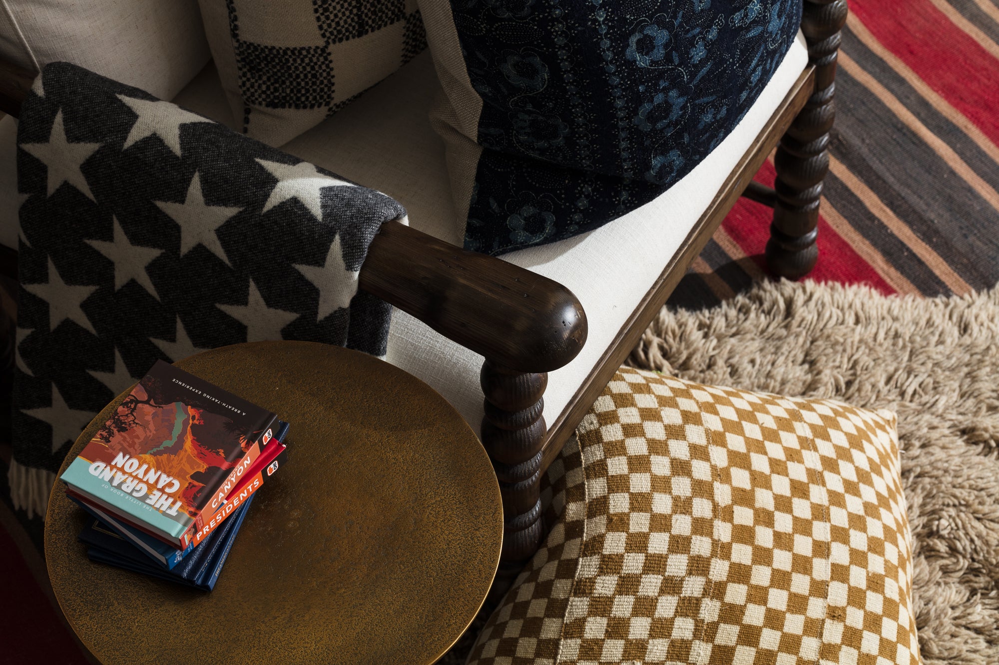 Cozy reading nook featuring a wooden-armchair with star-patterned blanket, a stack of books including "The Grand Canyon" on a round, bronze side table, and a checkered pillow on a plush, shaggy rug with striped carpet accent.