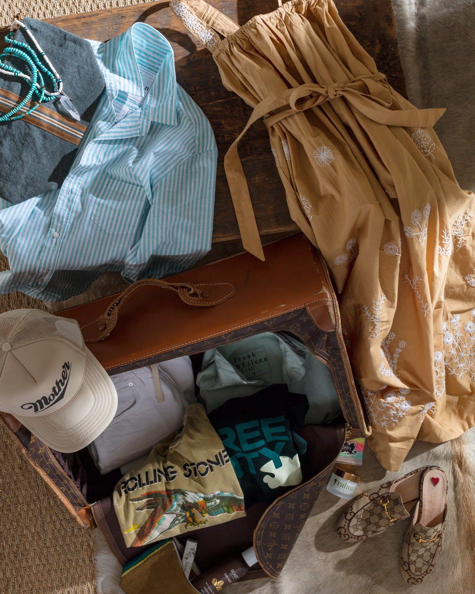 A travel suitcase is open and filled with folded clothes, including a cream-colored hat. Surrounding the suitcase are a light blue striped shirt with turquoise necklace, a beige maxi dress with embroidery, and a pair of patterned slip-on shoes.