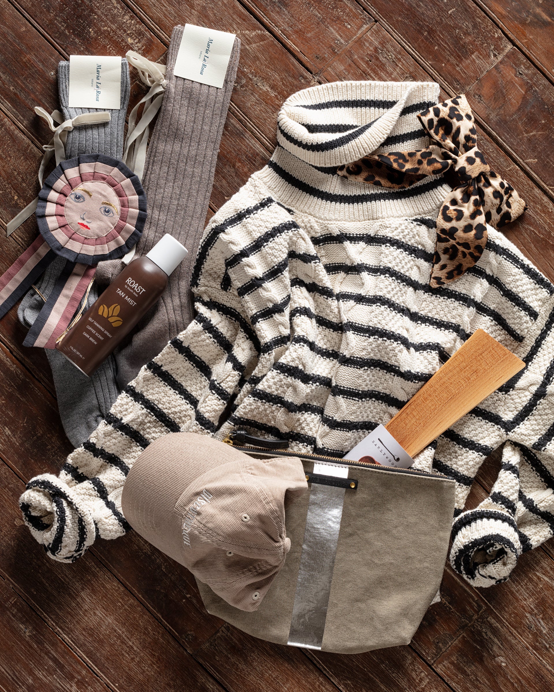 A striped sweater with a leopard print bow, gray socks, a doll decoration, a bottle of essence, a beige cap, and a small bag are arranged on a wooden floor.