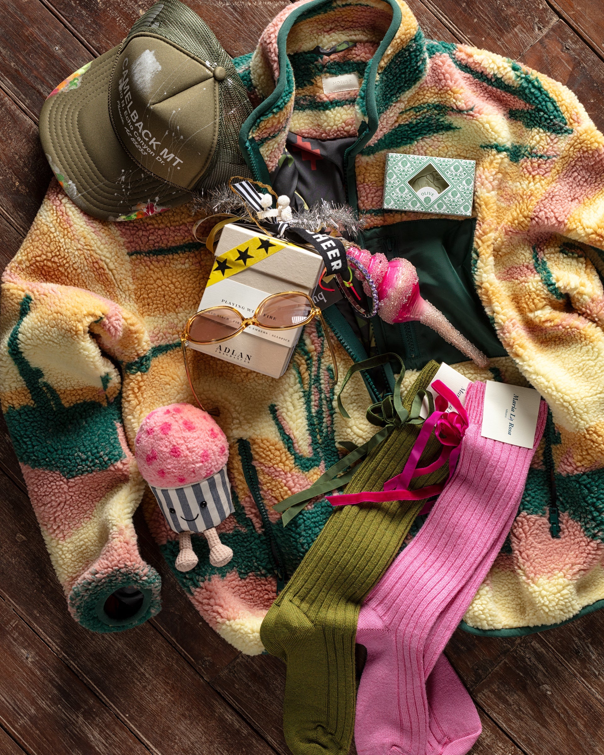 A colorful fleece jacket, green cap, perfume, sunglasses, pink and green socks, a makeup palette, and a pink plush toy arranged on a wooden surface.