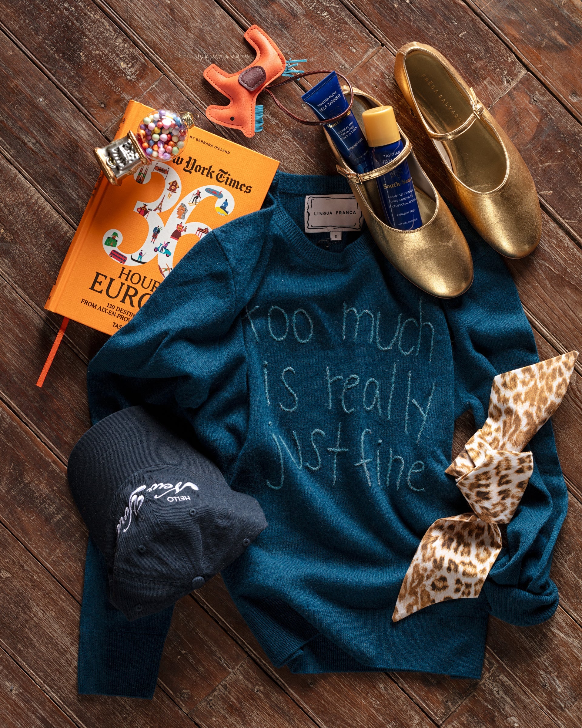 Flat lay of various items: a teal sweater with "too much is really just fine," golden shoes, a New York Times book, a bow, a toy, a black cap, and skincare products on a wooden floor.