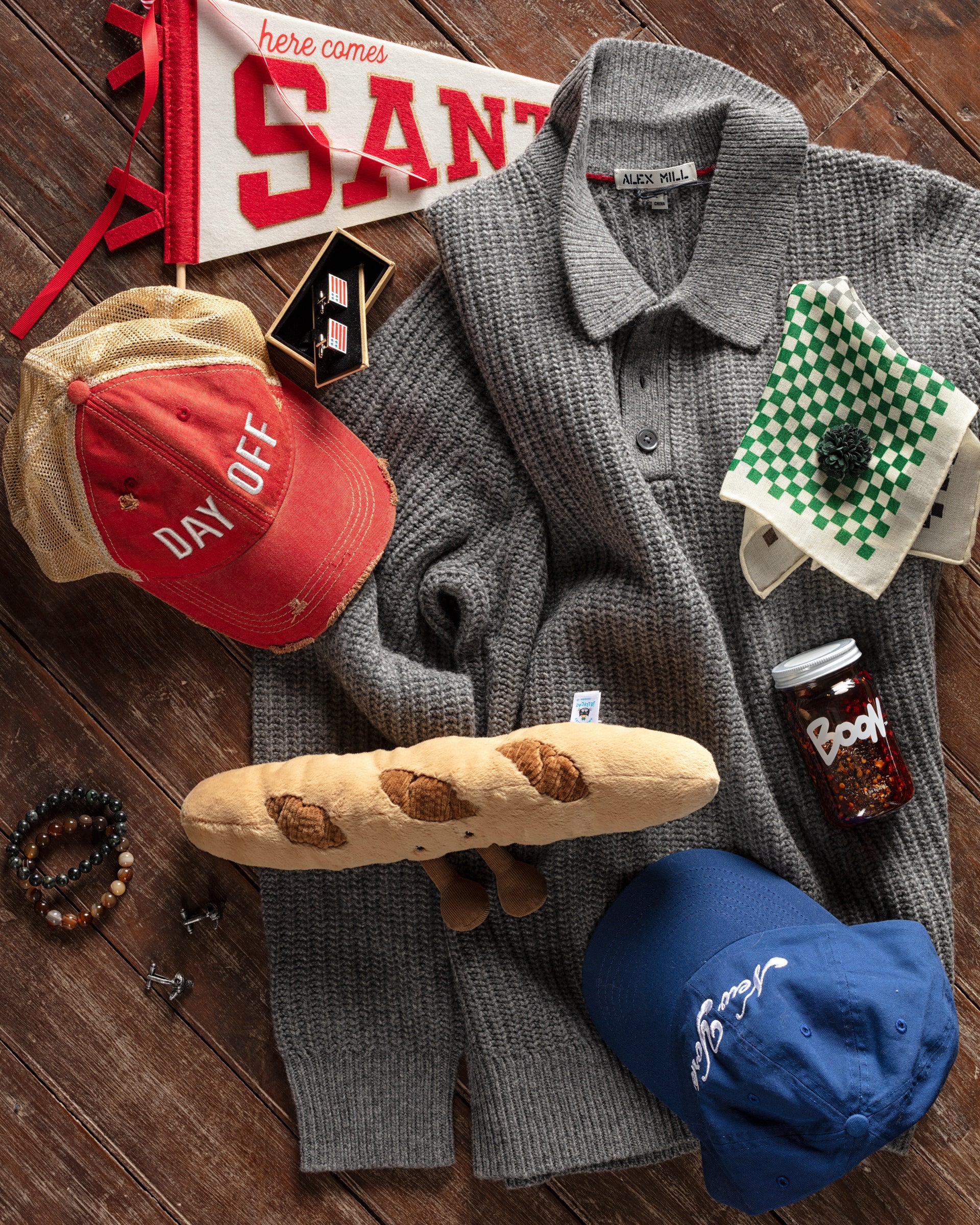 A collection of assorted items on a wooden surface, including sweaters, caps labeled "Day Off" and "Boon," a small flag, a stuffed baguette, bracelets, and checkered cloths.