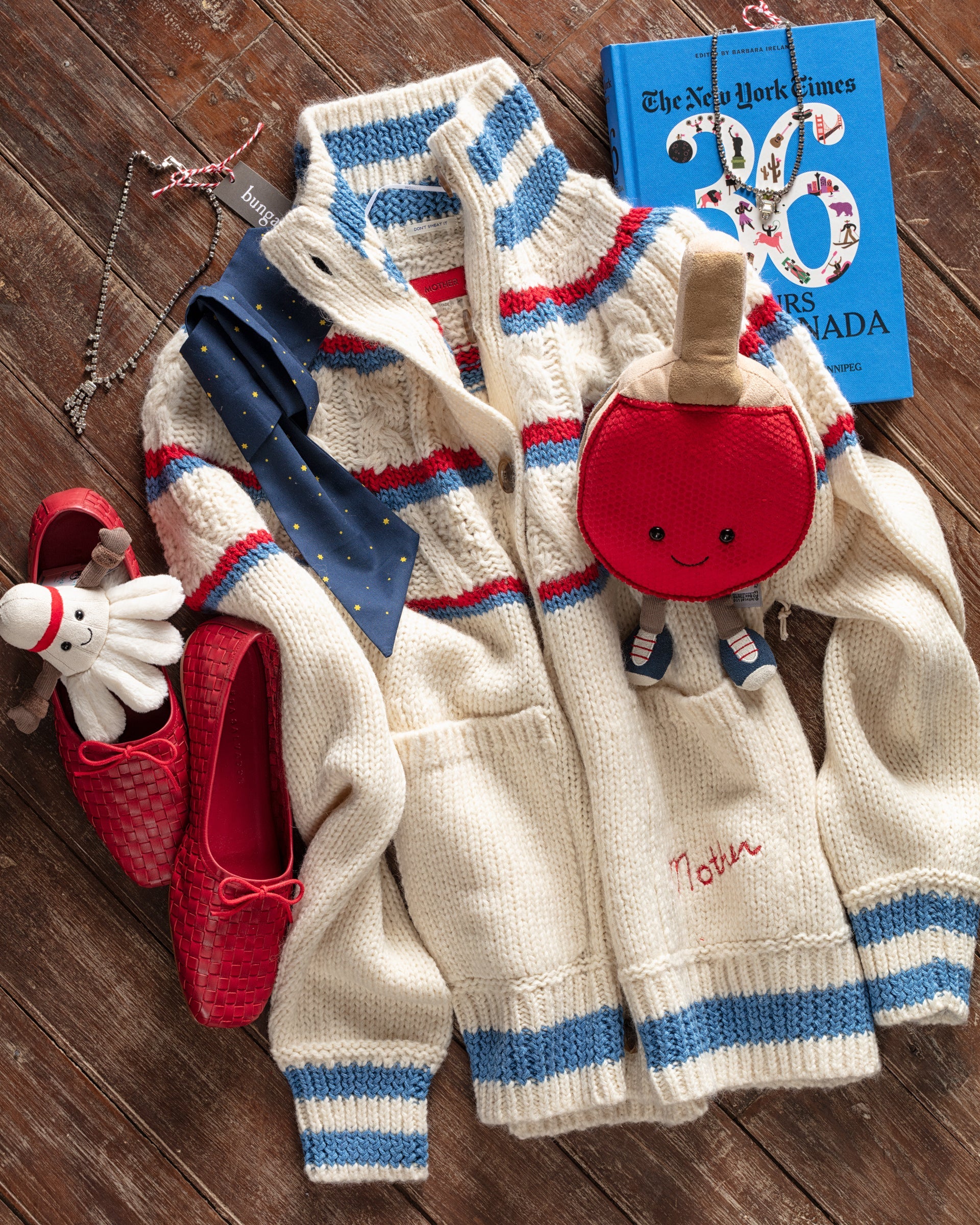 White knit sweater with red and blue stripes, alongside a blue tie, red ballet flats, plush octopus, red apple purse, and New York Times bestseller book. All arranged on a wooden surface.