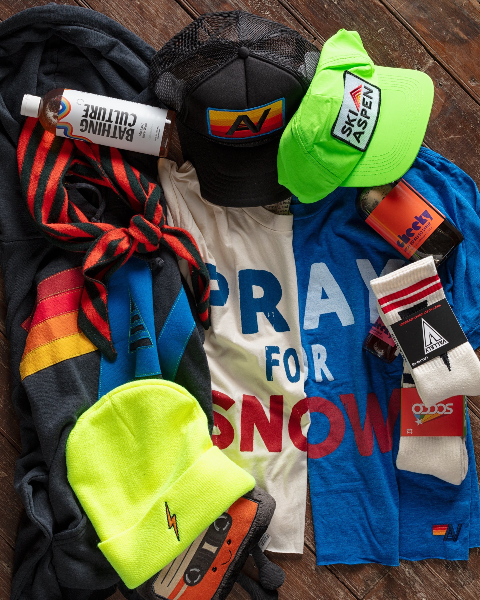 A collection of ski-themed clothing and accessories, including neon yellow hats, graphic T-shirts reading "Pray for Snow" and "Ski Aspen," a black netted cap, socks, a red-striped tie, and a bottle.