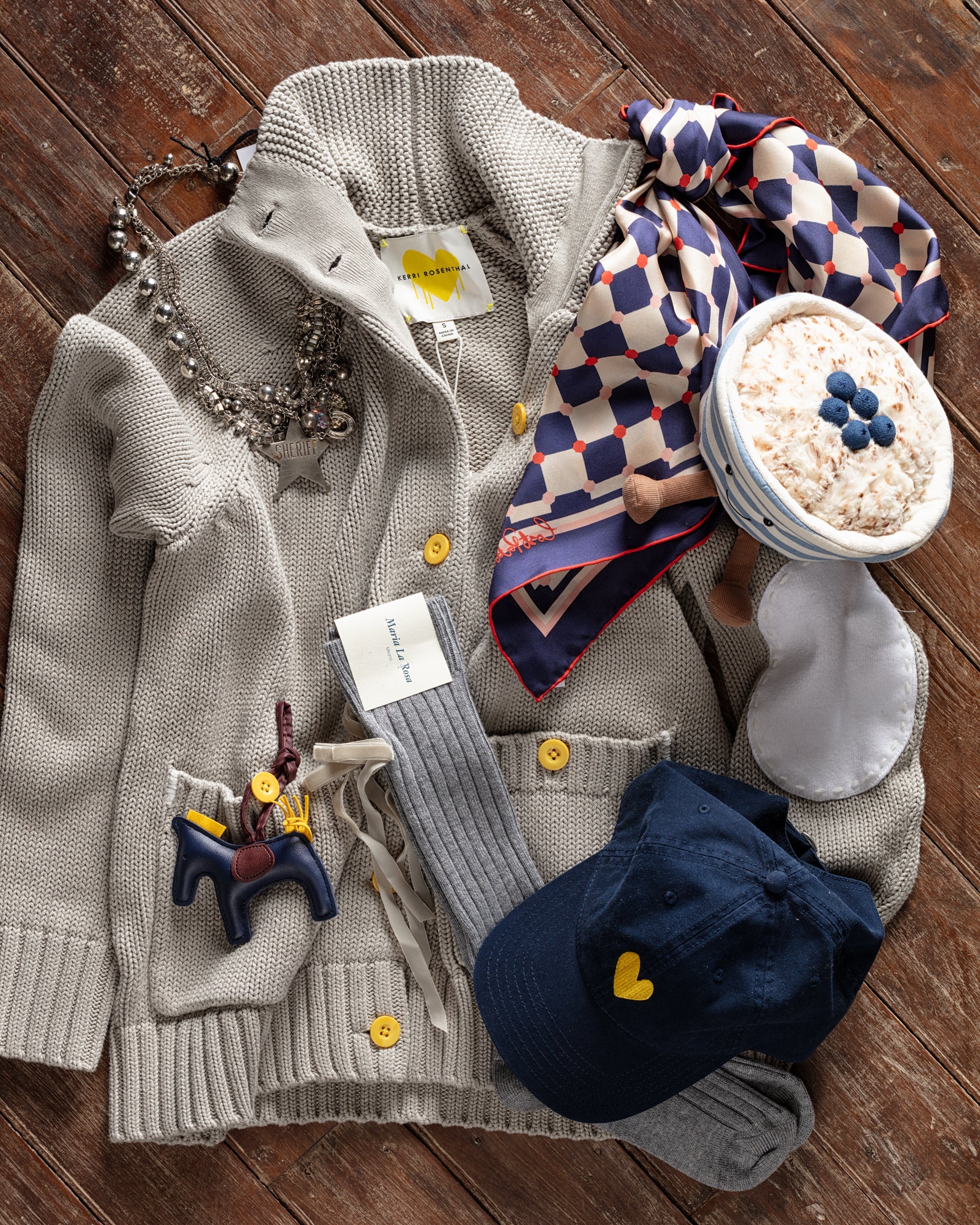 A beige cardigan with a necklace, patterned scarf, and a blue cap are laid on a wooden surface. Nearby are a bowl of oatmeal with blueberries, a sleep mask, and a gray belt with a tag.