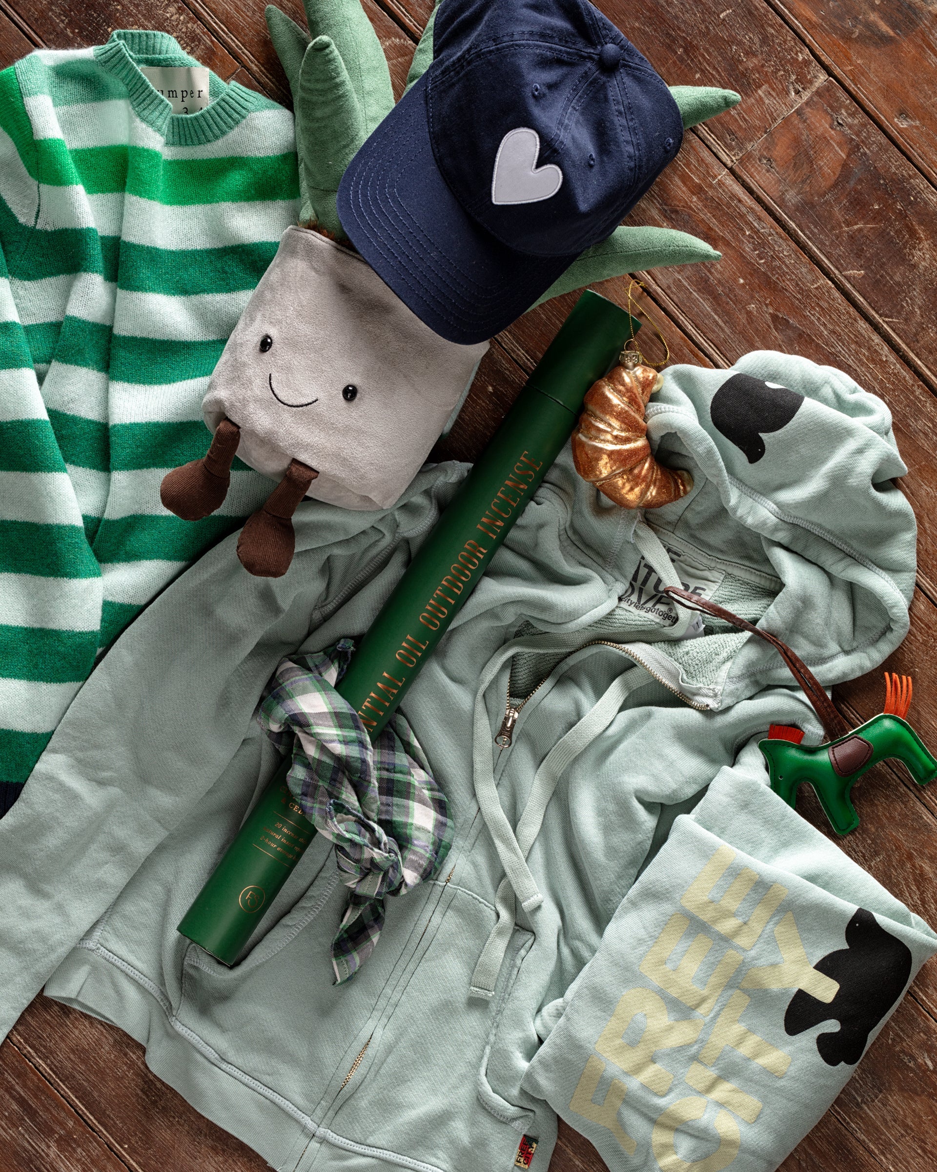 A green-themed flat lay featuring a striped sweater, plush toy, navy cap with a heart, hoodie, shirt, checkered tie, dinosaur figure, and a green tube on a wooden floor.