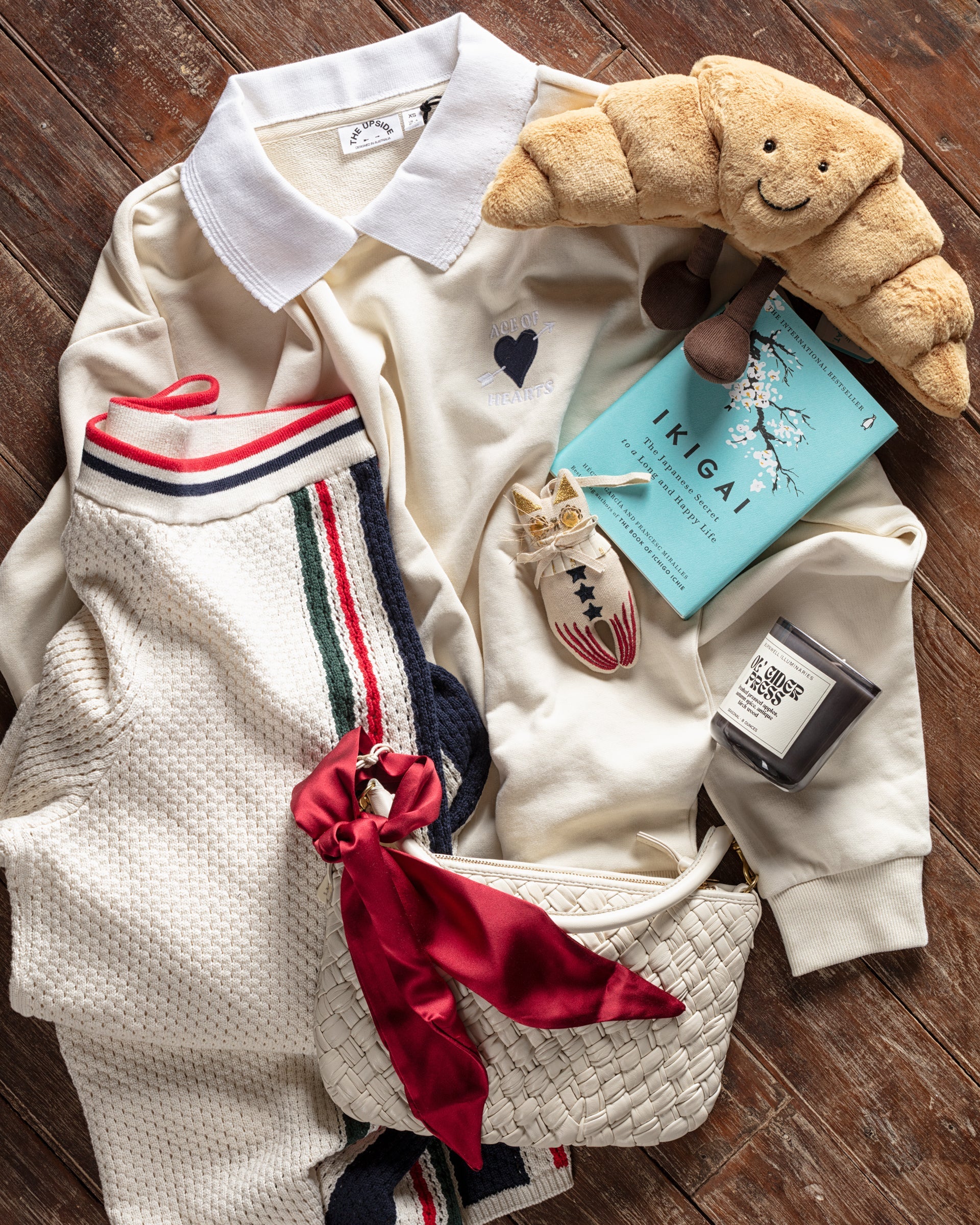 A cozy flat lay featuring a cream sweater, striped scarf, plush croissant toy, "Ikigai" book, and a candle. A white woven bag with a red ribbon completes the warm, textured arrangement on wooden flooring.