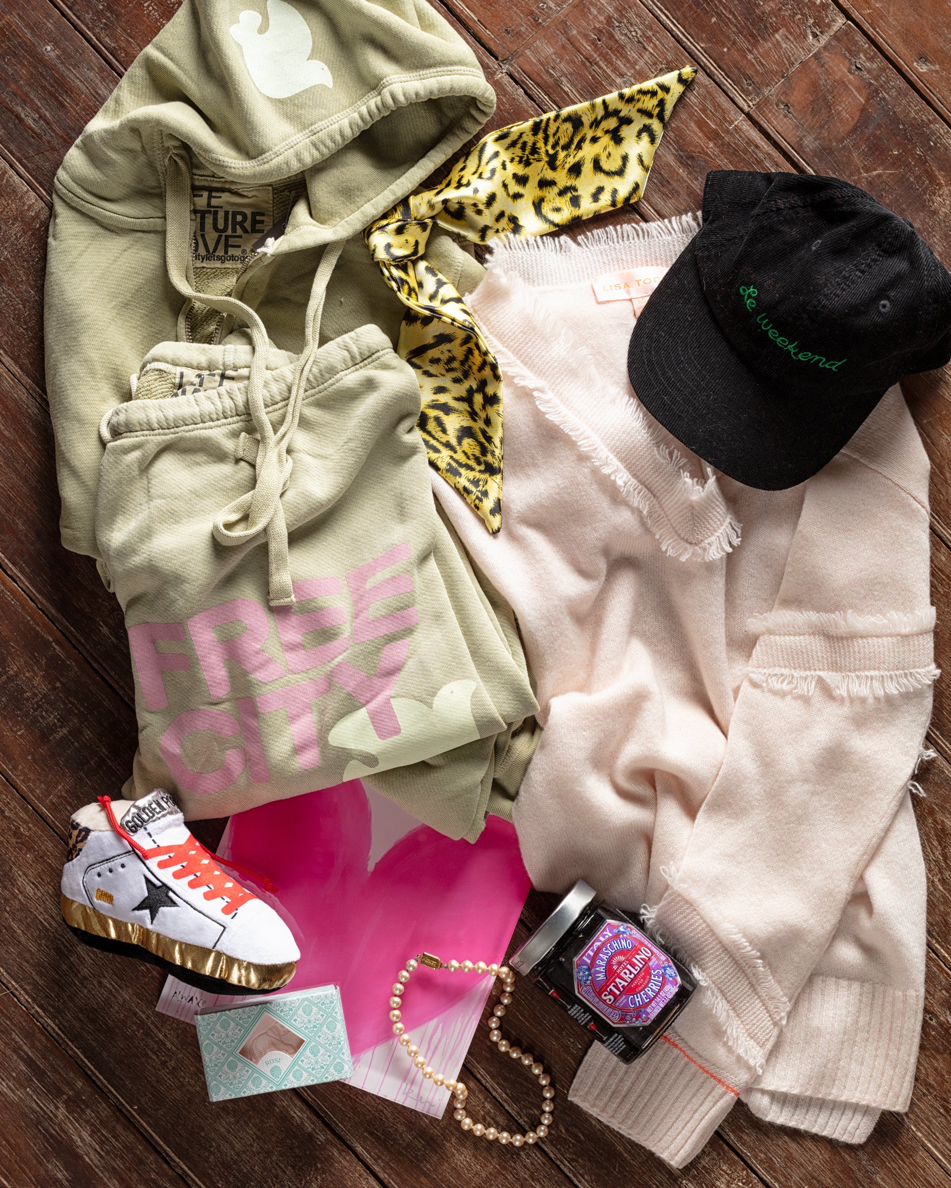 Clothing and accessories arranged on a wooden surface: green sweatshirt and pants, beige sweater, leopard print scarf, black cap, patterned sneakers, a skincare jar, pearl necklace, and a postcard.