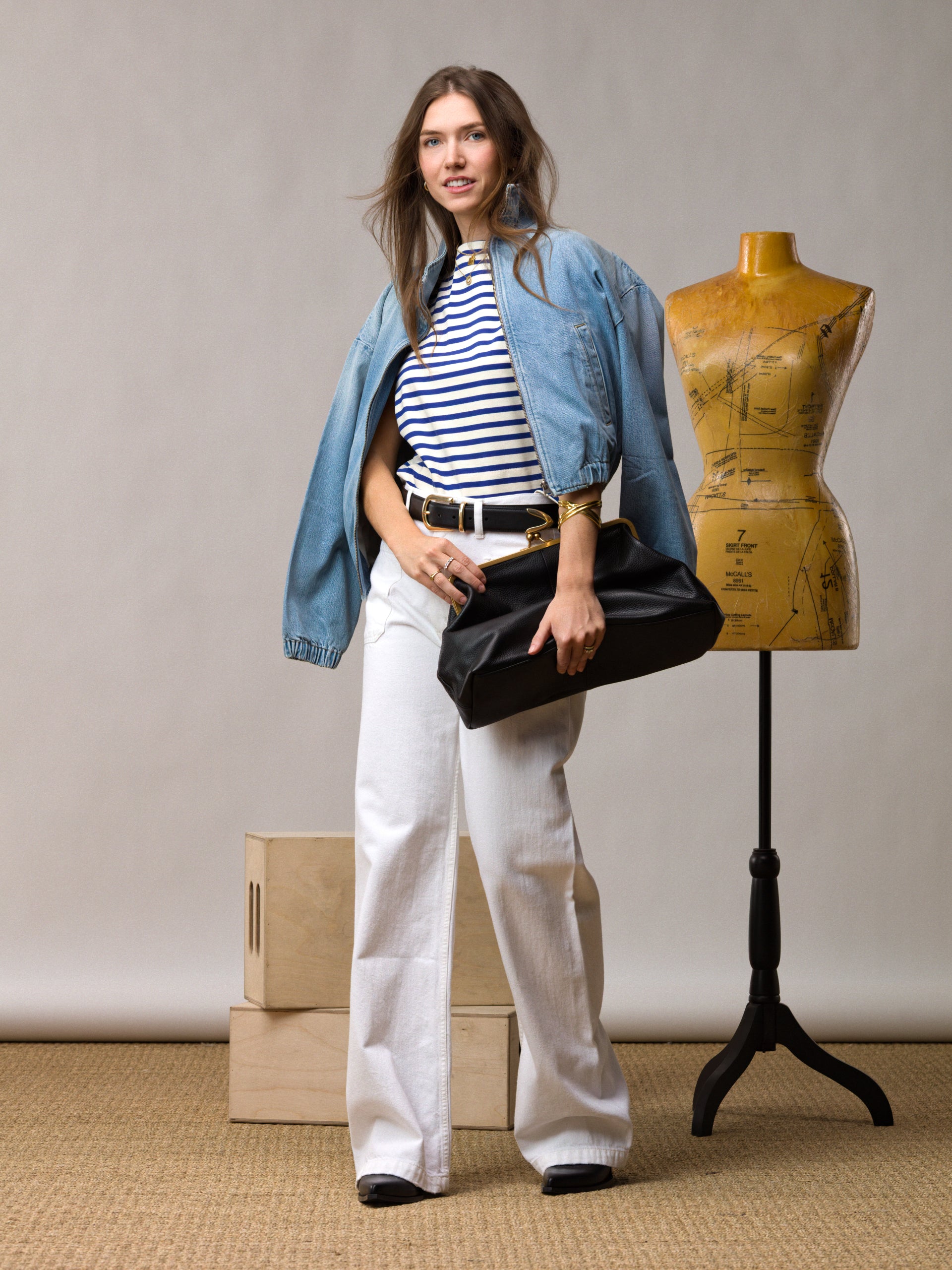 A woman wearing a striped shirt, denim jacket, and white pants holds a black clutch. She stands beside a dress form and wooden boxes against a neutral background.