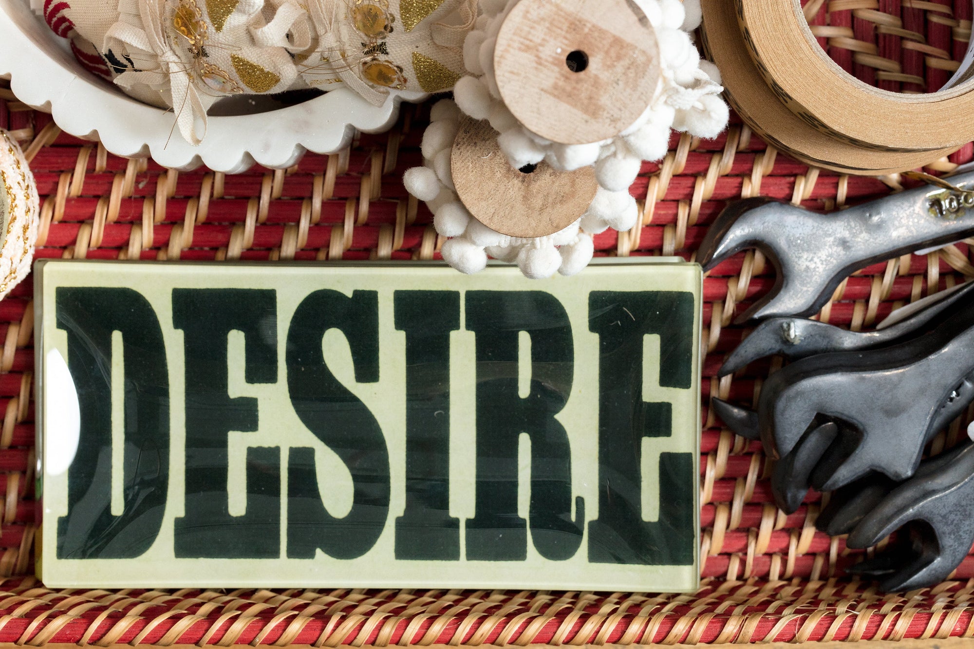 A woven surface holds a sign with the word "DESIRE" in bold letters, surrounded by crafting materials like ribbon, wooden spools, and metal wrenches.