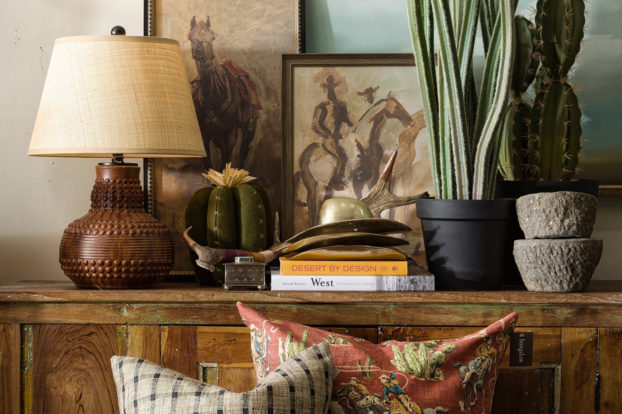 A rustic wooden table displays a woven lamp, stacked books, potted cacti, and framed Western art, with patterned pillows arranged beneath.