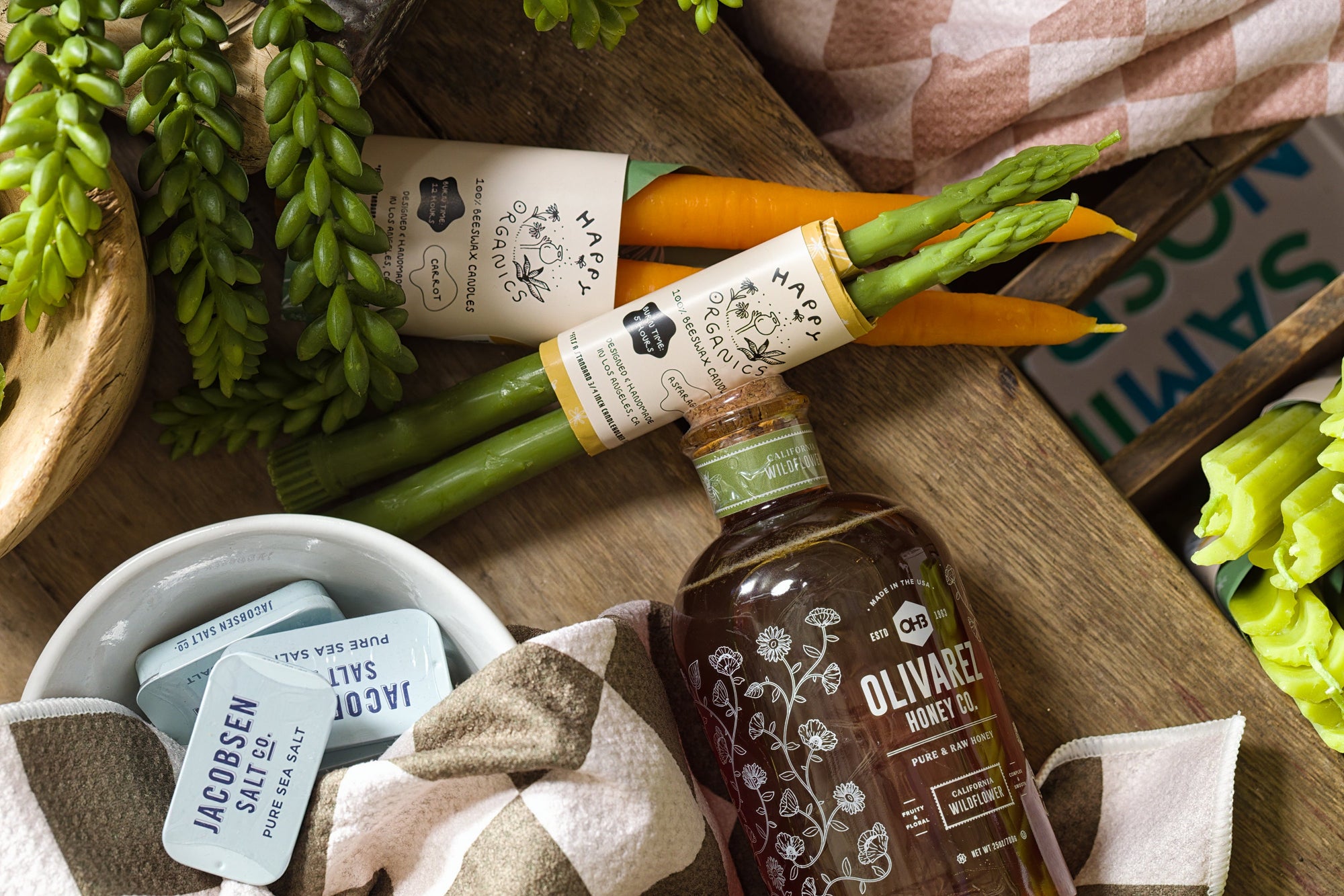 A bottle of organic olive oil, a bundle of fresh asparagus and carrots, sea salt packets in a bowl, and green succulents arranged on a rustic wooden surface with a checkered cloth.
