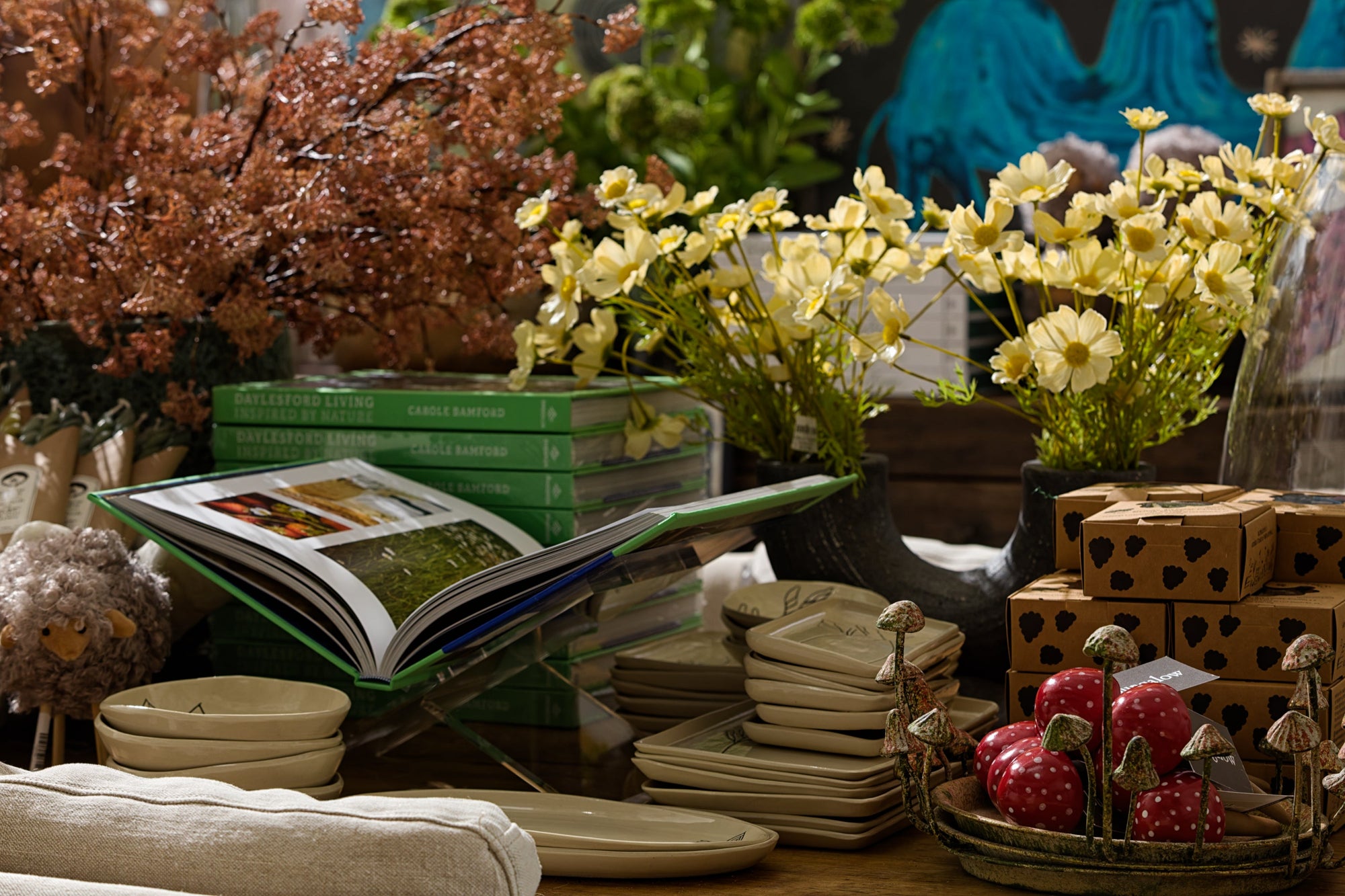A cozy display with stacked books, an open book on a stand, ceramic plates, heart-shaped dishes, fake mushrooms, wrapped gifts, and decorative flowers in vases.