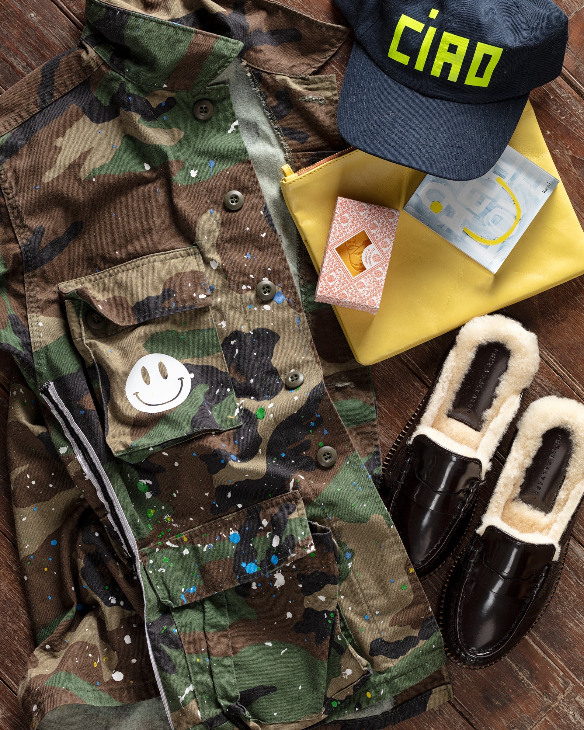 A camouflage jacket with paint splatters and a smiley patch, a navy cap with "CIAO," a yellow envelope, a pouch of facial tissues, and a pair of black fur-lined loafers are displayed.