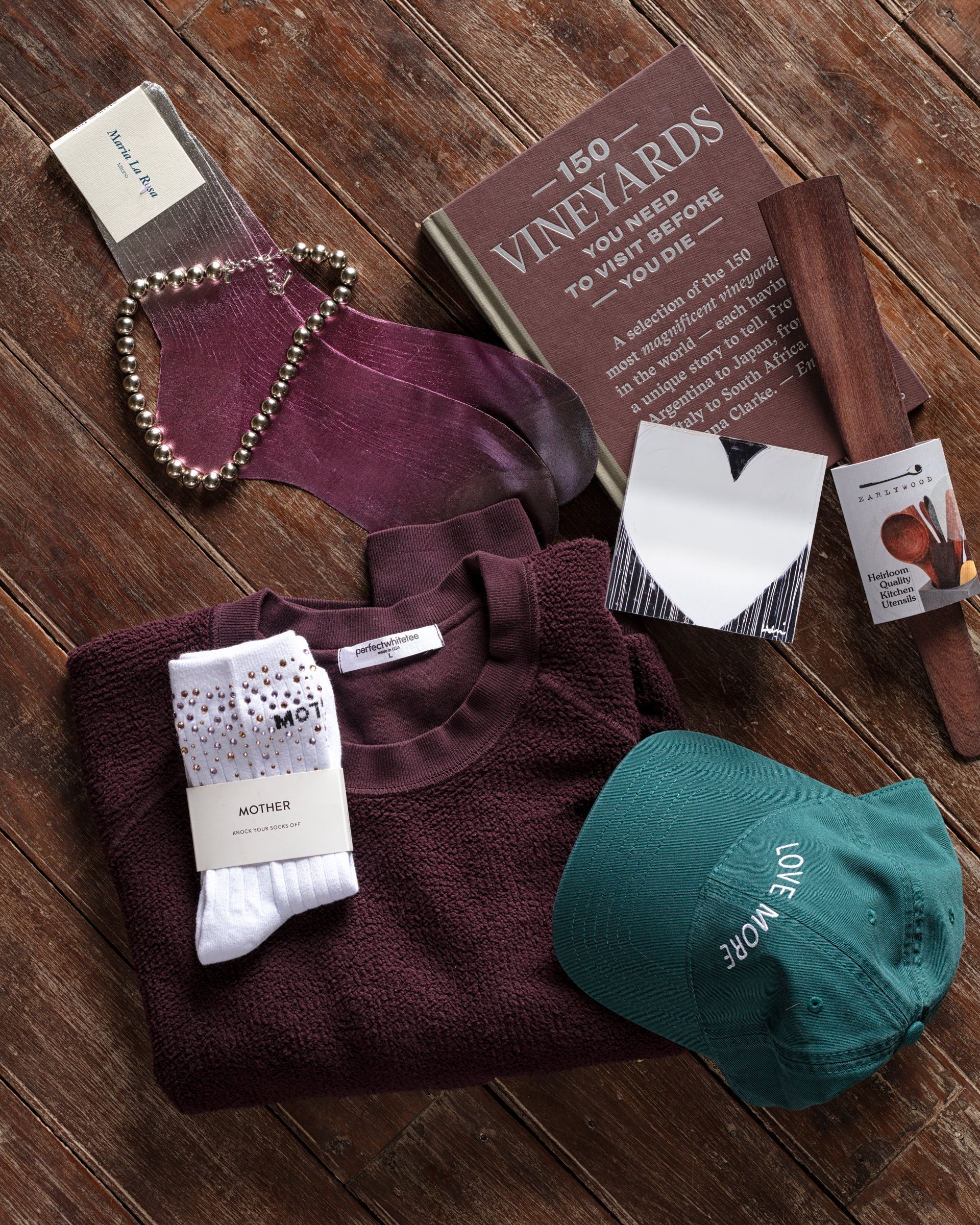 A flat lay featuring a maroon sweater, a green cap with "LOVE MORE" text, a book titled "150 Vineyards," purple socks, white socks, a necklace, a dark wooden object, and a scented ornament on a wooden surface.