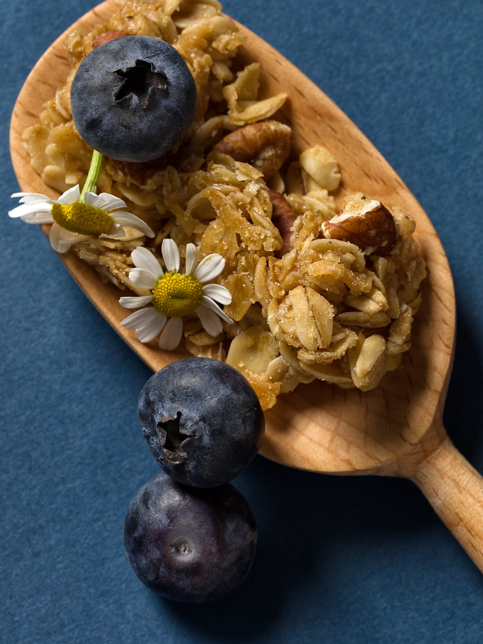 A bowl of granola with blueberries and nuts on a wooden board, garnished with small flowers. Blueberries and a blue-patterned napkin are placed nearby.
