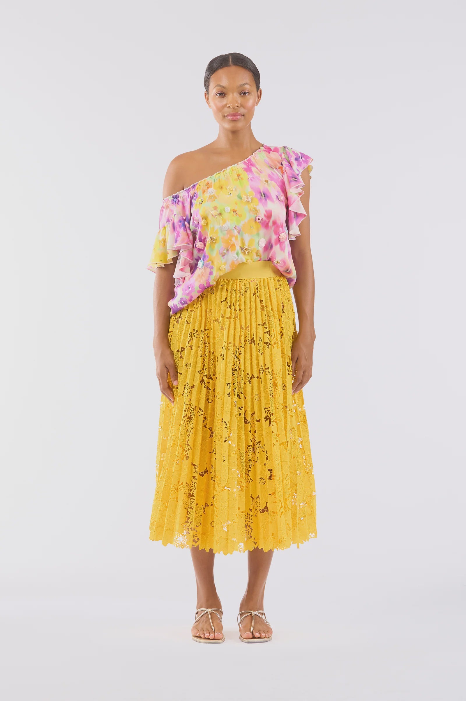 Against a white background, a person with short hair wears an off-the-shoulder top adorned with pink and yellow patterns and pairs it with Le Superbe's Sunflower Pleated Skirt, which features floral lace details in golden yellow. They complete the look with sandals.