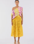 Against a white background, a person with short hair wears an off-the-shoulder top adorned with pink and yellow patterns and pairs it with Le Superbe's Sunflower Pleated Skirt, which features floral lace details in golden yellow. They complete the look with sandals.