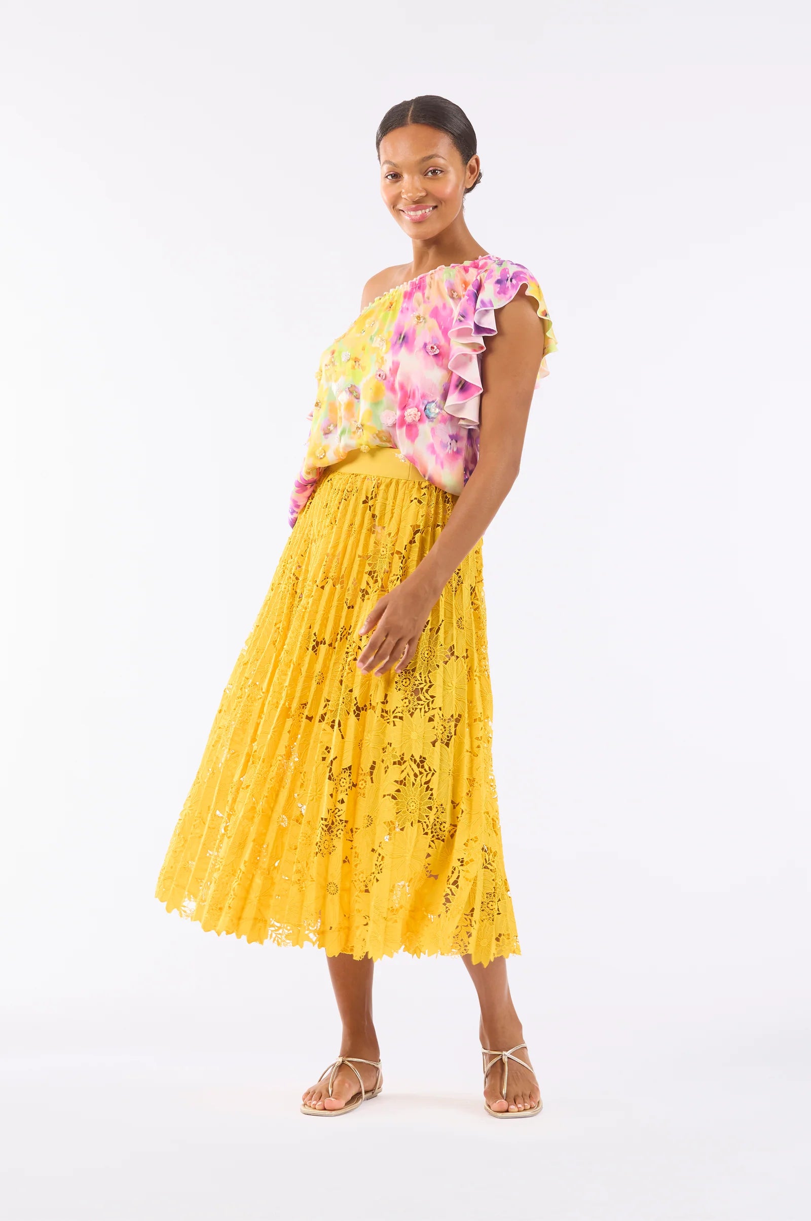 A woman models a colorful one-shoulder floral top and the "Sunflower Pleated Skirt" by Le Superbe against a white background. She pairs it with white strappy sandals, smiling radiantly with hands relaxed at her side. The skirt offers comfort with its elegant elastic waistband.