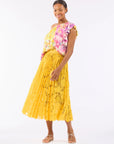 A woman models a colorful one-shoulder floral top and the "Sunflower Pleated Skirt" by Le Superbe against a white background. She pairs it with white strappy sandals, smiling radiantly with hands relaxed at her side. The skirt offers comfort with its elegant elastic waistband.