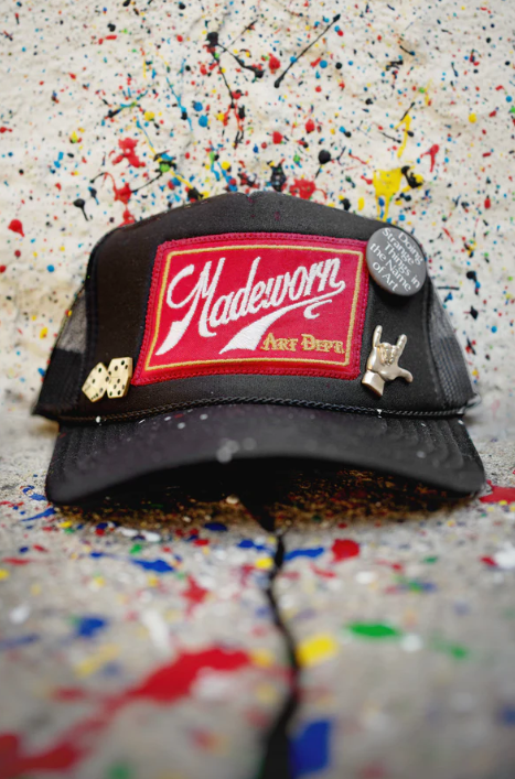 The MadeWorn Hat by Made Worn is a black trucker cap with a custom "Madeworn Art Dept" front patch, limited edition pins (dice, hand sign, and “The best things in life aren't things”), an adjustable back strap, and a paint-splattered backdrop.