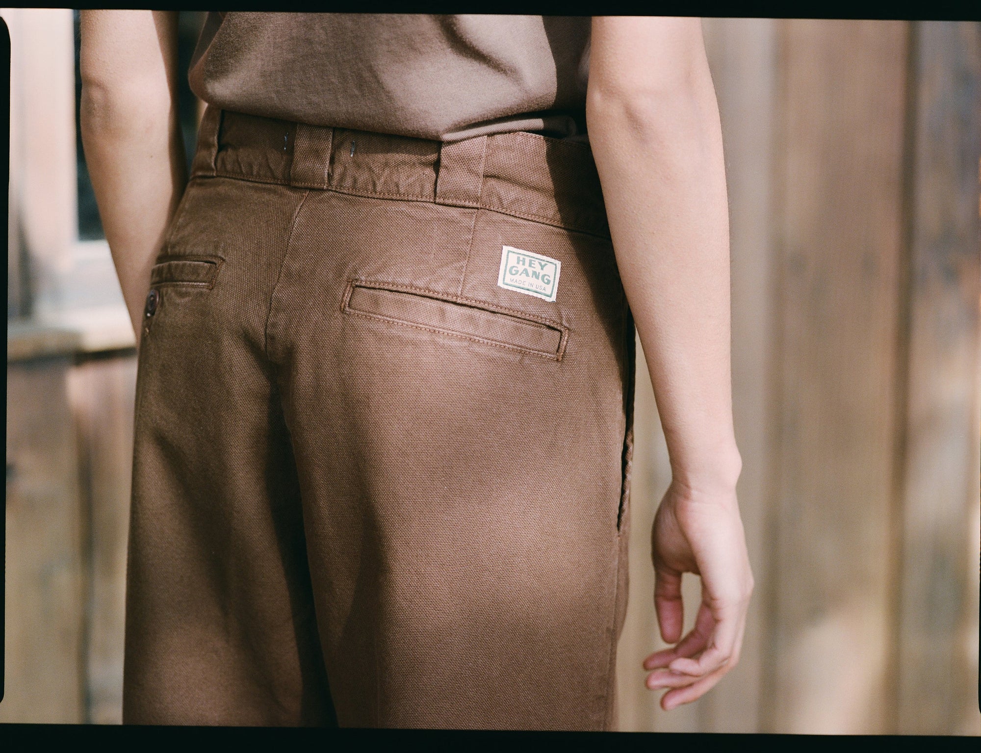 Someone wearing Hey Gang’s The Work Pant in brown cotton canvas—with a branded patch on the back pocket—stands by a wooden wall, their right arm relaxed at their side.