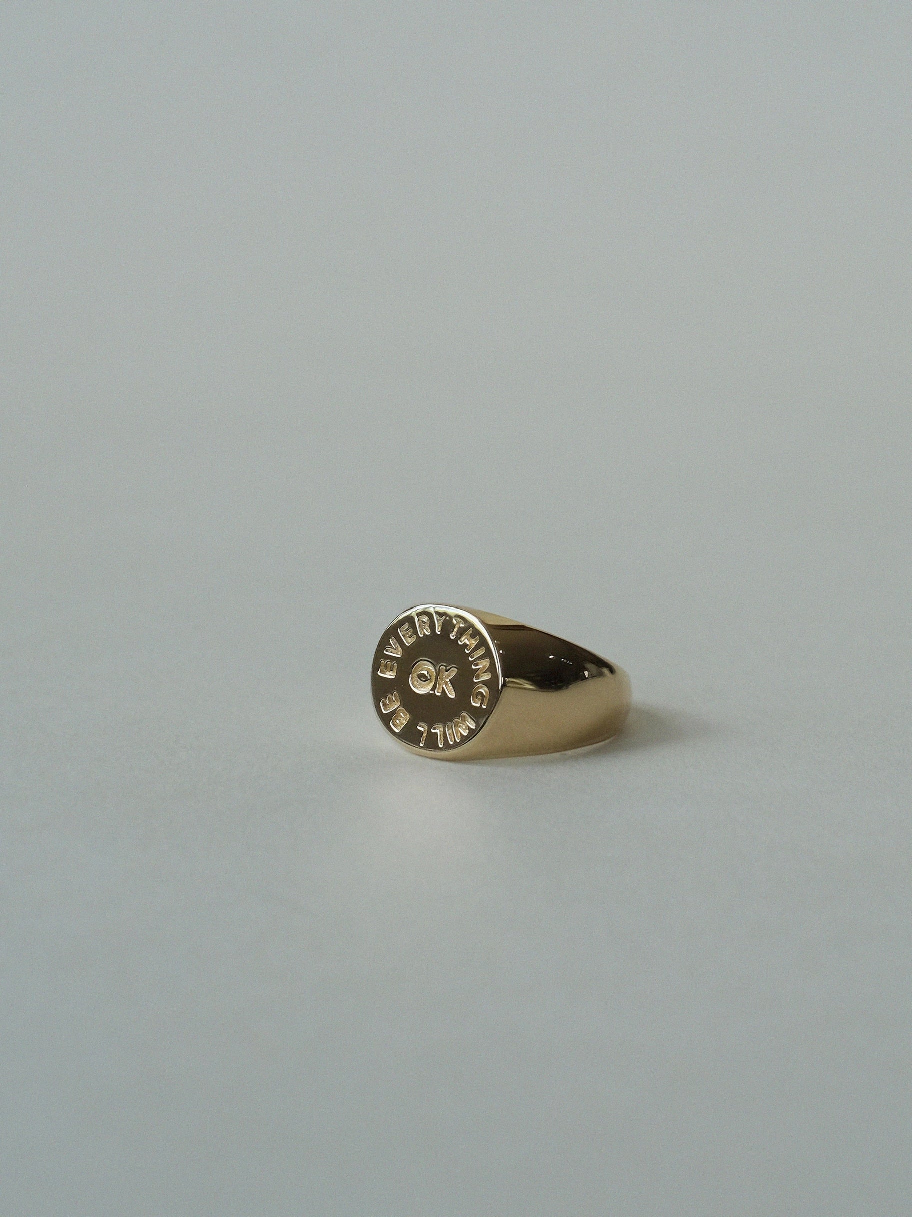 The Faire "Everything Will Be OK Ring" is a gold-plated signet, engraved with "ALL THINGS EVENTUALLY" in a circular pattern around a small central symbol, shown resting on a light gray surface.