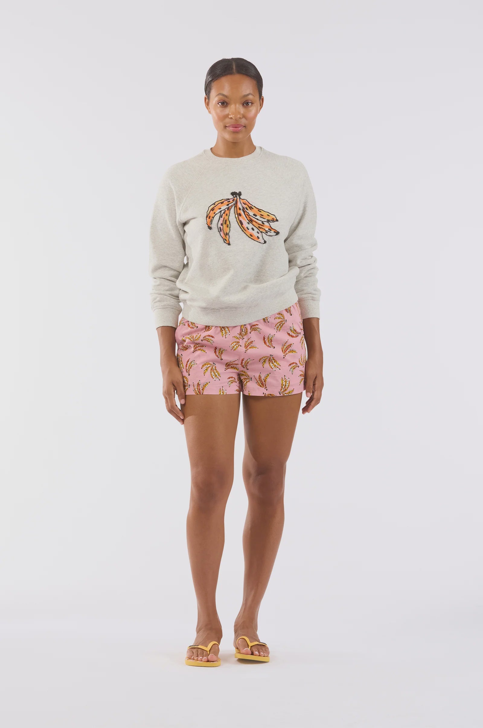 A person stands against a plain background wearing Le Superbe's Beaded Bananas Sweatshirt in ultra-soft fabric, paired with pink banana-patterned shorts and yellow flip-flops.