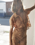 A woman poses in a doorway, sunlight highlighting her Le Superbe Suntan Camp Shirt. With one arm raised to the doorframe, she stands against an urban street backdrop, showcasing the shirt’s effortless style.
