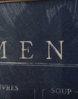 Close-up of a worn, textured Menu Art menu cover from a Scottsdale Arizona bungalow, with the word "MENU" embossed in gold lettering, showing signs of aging and scratches. Brand: Mercana