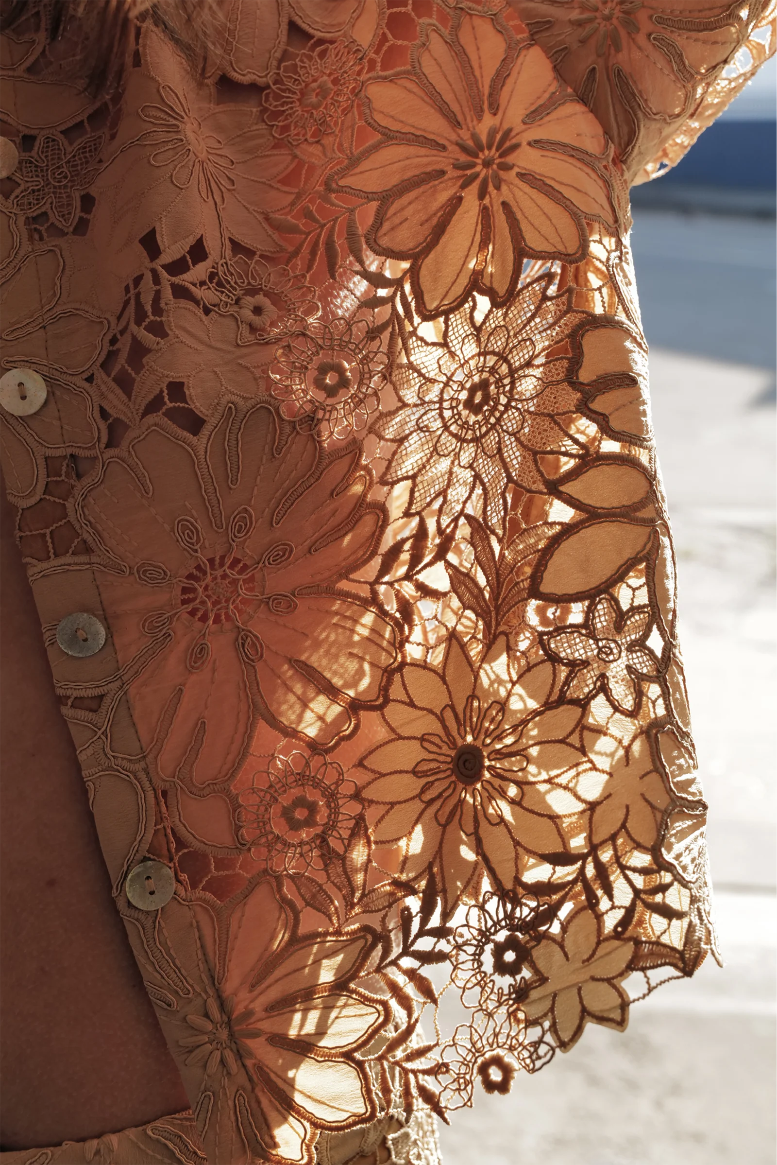 Close-up of Le Superbe's Suntan Camp Shirt, a sheer peach blouse with intricate floral lace, scalloped hem, and button details. Sunlight highlights its delicate patterns and casts soft shadows through the fabric.