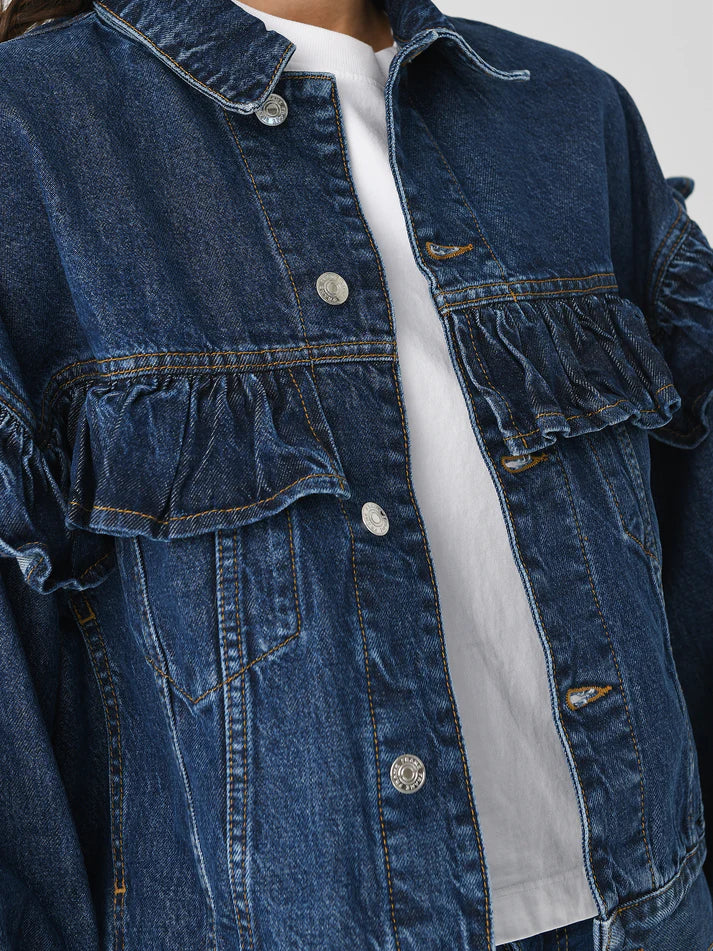 A close-up highlights a person donning FRAME's Ruffle Jacket, a classic fit denim piece with ruffle detail across the chest and shoulders, adorned with metallic buttons. It's layered over a white top against an off-white backdrop.