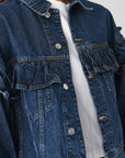 A close-up highlights a person donning FRAME's Ruffle Jacket, a classic fit denim piece with ruffle detail across the chest and shoulders, adorned with metallic buttons. It's layered over a white top against an off-white backdrop.