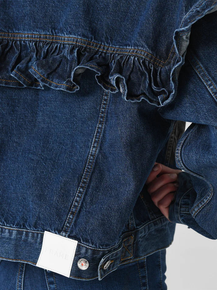 A close-up of FRAME's Ruffle Jacket with visible stitching, ruffle detail on back and sleeves, a white label at the bottom, held by a person in matching jeans.