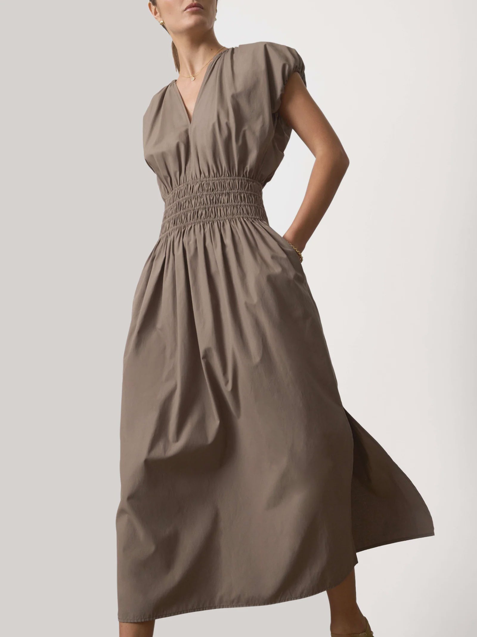 A woman wears the BROCHU WALKER MINA DRESS, a taupe midi dress with a shirred V neckline, short sleeves, smocked waist, full skirt, pockets, and an elegantly relaxed fit. The background is plain and light-colored.