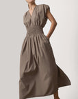 A woman wears the BROCHU WALKER MINA DRESS, a taupe midi dress with a shirred V neckline, short sleeves, smocked waist, full skirt, pockets, and an elegantly relaxed fit. The background is plain and light-colored.