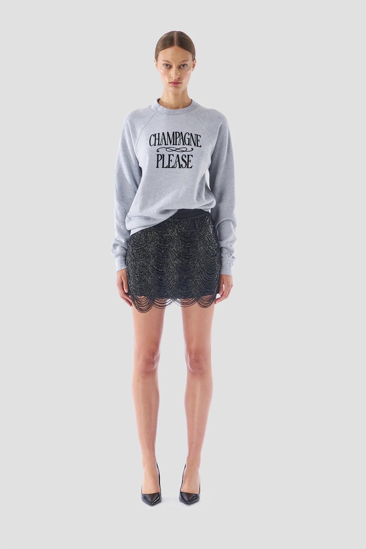 A woman poses against a plain backdrop, showcasing the "Champagne Please" sweater by Le Superbe in gray. She complements it with a black glittery skirt that features a scalloped hemline and finishes her outfit with black high heels. Her hair is styled in an updo.