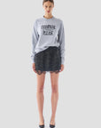 A woman poses against a plain backdrop, showcasing the "Champagne Please" sweater by Le Superbe in gray. She complements it with a black glittery skirt that features a scalloped hemline and finishes her outfit with black high heels. Her hair is styled in an updo.